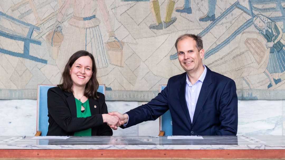 To personer i formell antrekk håndhilser over et bord med papirer. Et stort veggmaleri med figurer er i bakgrunnen.