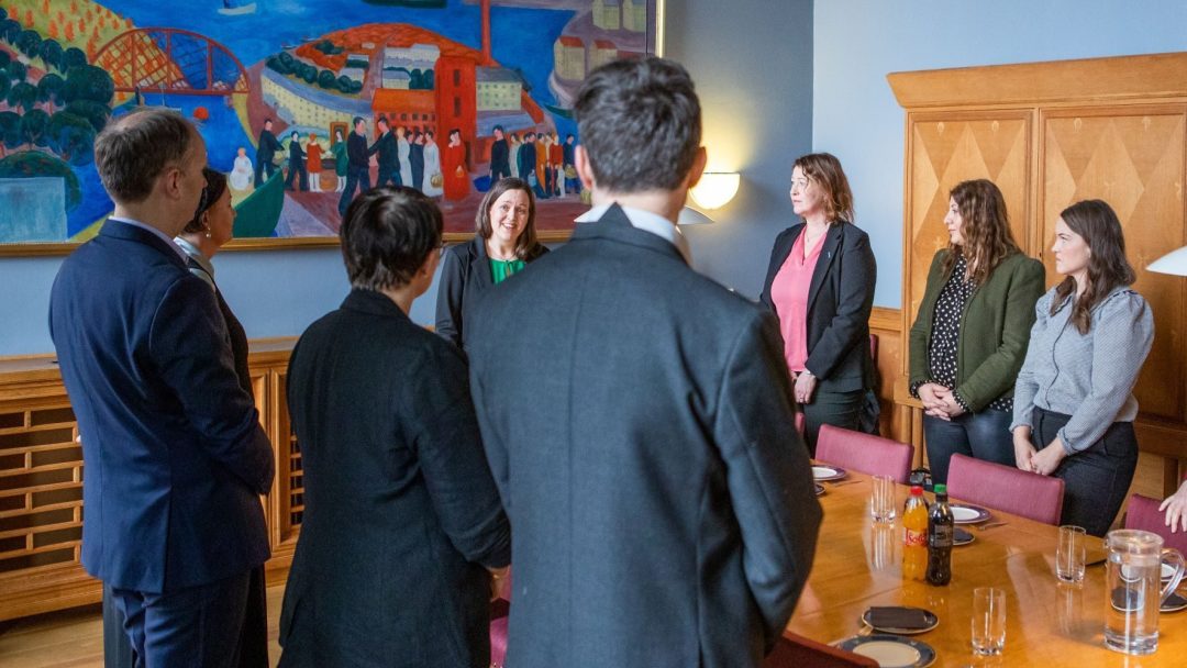 En gruppe mennesker i formell antrekk står rundt et trebord i kontormiljø og snakker. Et fargerikt maleri er på veggen i bakgrunnen.
