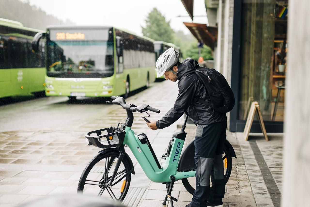 En person i regntøy justerer en blågrønn elektrisk sykkel merket «TIER» på et vått fortau, med grønne busser i bakgrunnen på en regnværsdag.