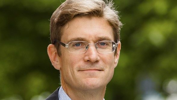 En mann i dress og briller står selvsikkert, med Klimagassregnskapet som subtilt påvirker det uskarpe grønne bakteppet.