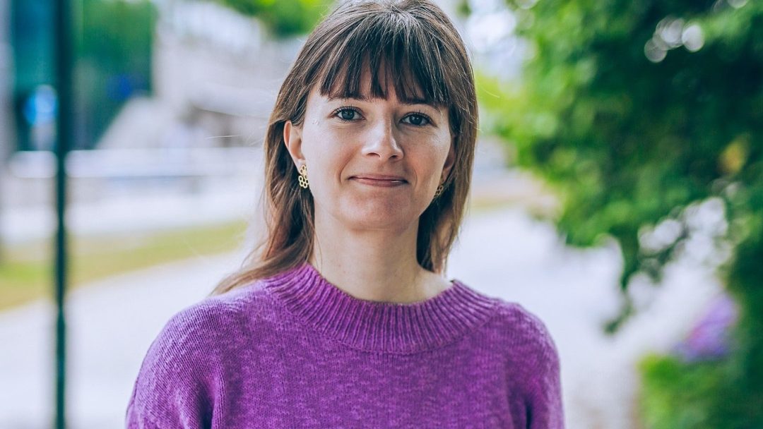 En person med langt hår iført en lilla genser står utendørs med grønt og uskarp bakgrunn.