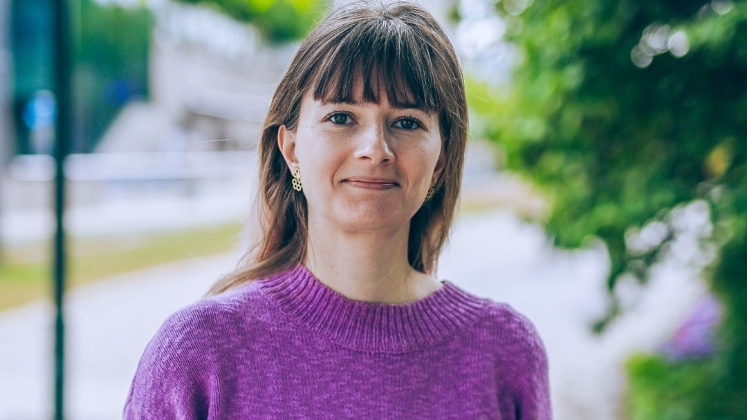 En person i lilla genser står utendørs med grønt i bakgrunnen, nær praktiske ladeplasser for elektriske kjøretøy.