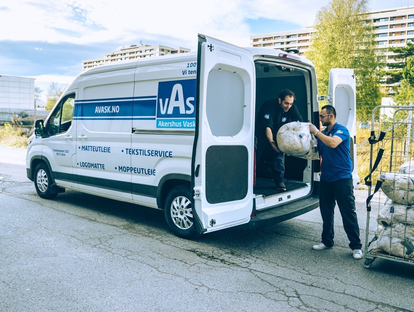 To menn laster en varebil med store sekker. Varebilen er hvit med et firmas merke på siden. Bygninger og trær er synlige i bakgrunnen.