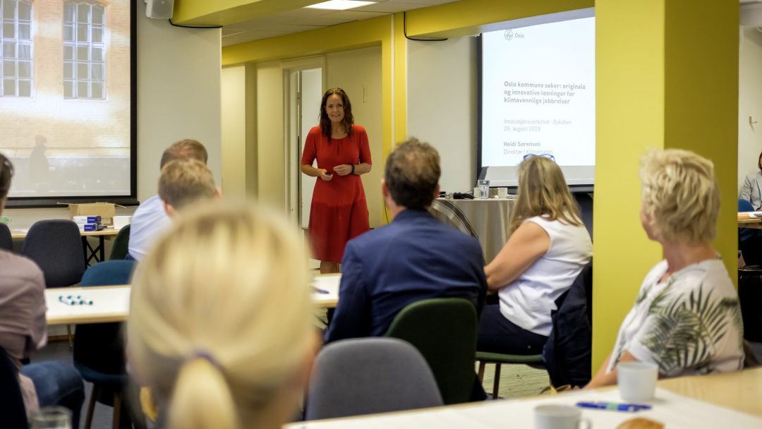 En kvinne i en rød kjole snakker til et publikum under en presentasjon i et sterkt opplyst rom. Deltakerne sitter ved bordene, vendt mot høyttaleren og en skjerm som viser et lysbilde.