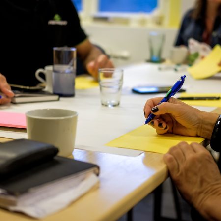 Folk rundt et bord som skriver på gult papir med penner, omgitt av notatbøker og krus.