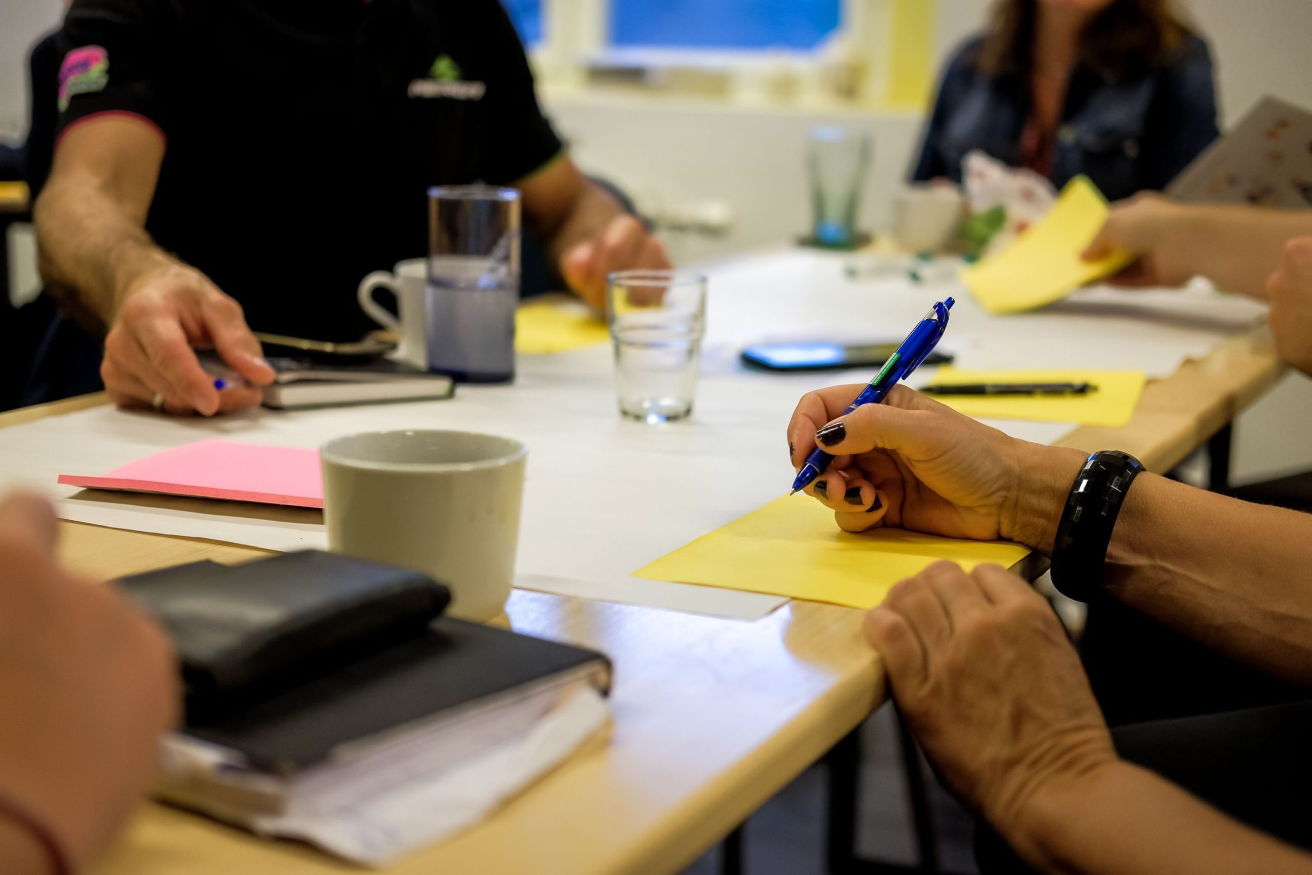 Folk rundt et bord som skriver på gult papir med penner, omgitt av notatbøker og krus.