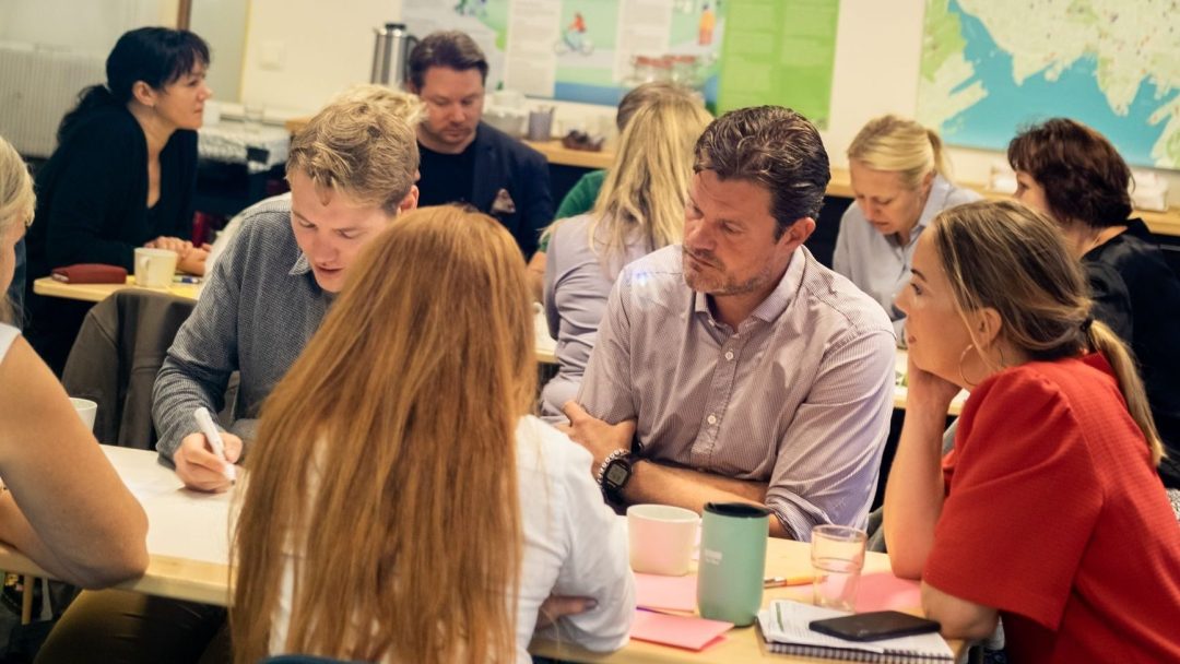 Folk som satt ved bord og diskuterte, med kart på veggen bak seg.