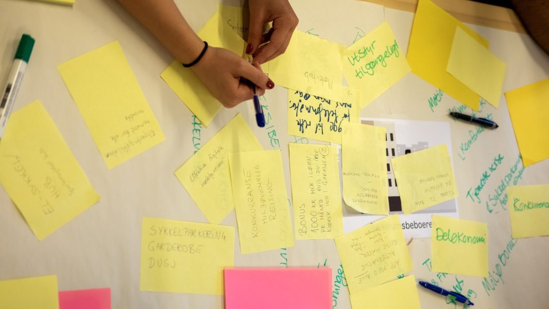 Person som skriver på klistrelapper spredt på et bord, med en hånd som holder en penn over en gul lapp. Ulike håndskrevne notater og markører er synlige.