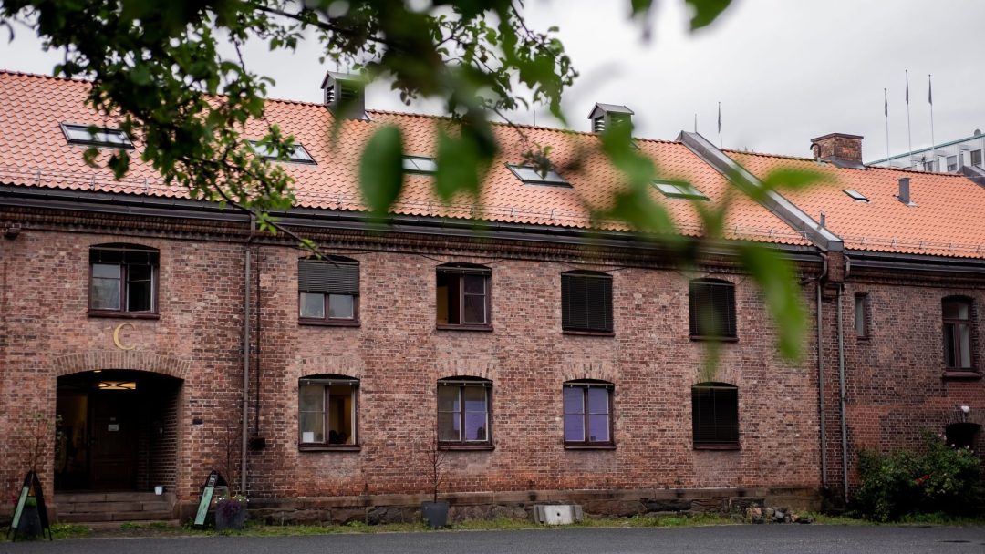 Murbygning med rødt tegltak og flere vinduer, delvis skjult av grønne blader i forgrunnen.