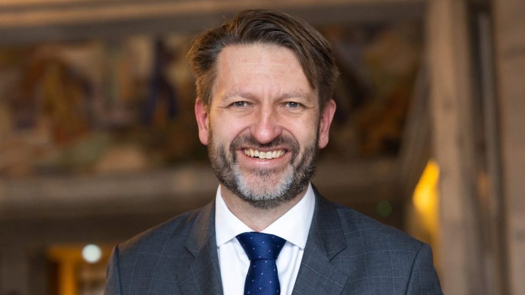 En smilende mann i dress og slips med skjegg står selvsikkert i en innendørs setting og holder et fraktdokument.