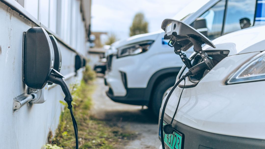 To elbiler med klimatilskudd kobles til veggmonterte ladere på en parkeringsplass, med en bil i forgrunnen og en annen i bakgrunnen.