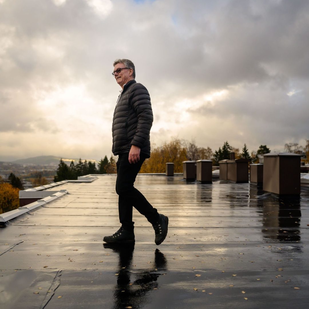 En person i mørk jakke og bukser går på et flatt tak, og ser på sameier mot et overskyet bakteppe, med bybildet og trærne som strekker seg utover.