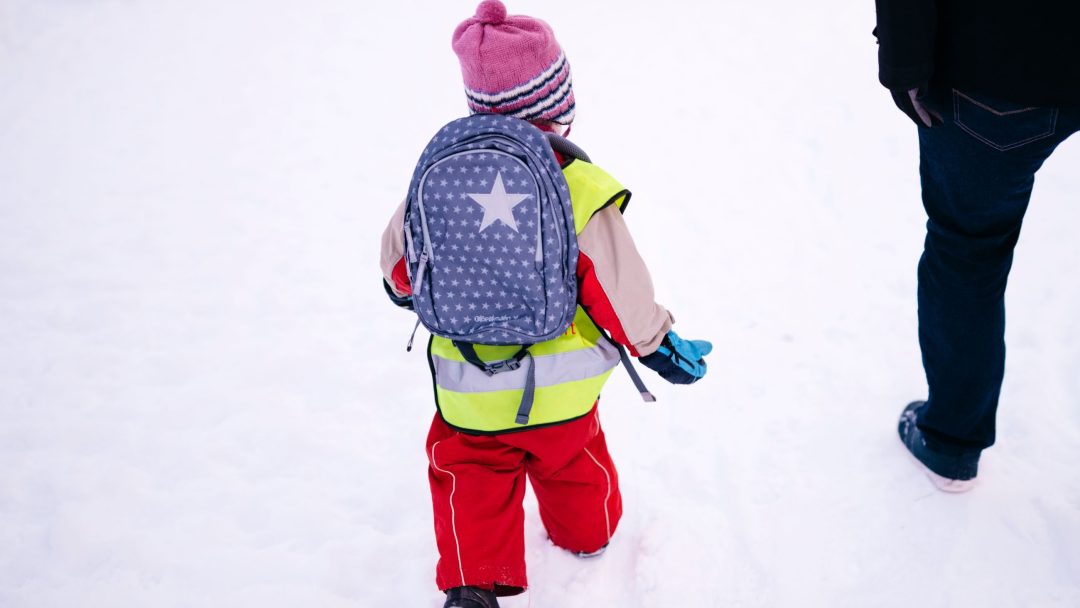 Et barn i røde snøbukser, gul vest og stripete lue, med en stjernemønstret ryggsekk som gir et snev av innfall, går i snøen ved siden av en voksen. Reisen deres gjennom vinterlandskapet understreker subtilt temaer fra en klimasårbarhetsanalyse.