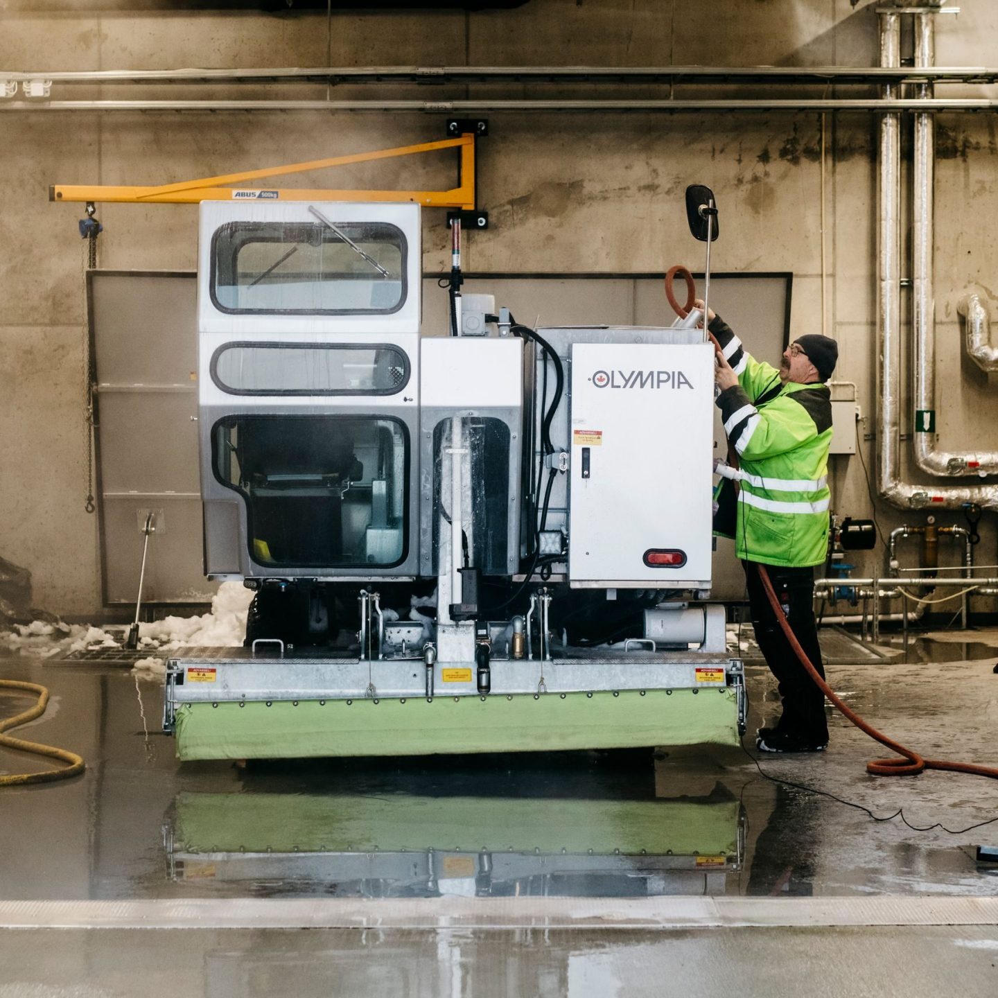 Person i synlig jakke som rengjør en isfornyingsmaskin med en slange i en industrigarasje.