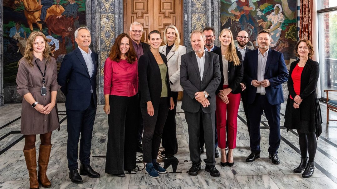 En gruppe på tolv personer, som står innendørs på et marmorgulv, og poserer for et bilde foran en dekorativ vegg.