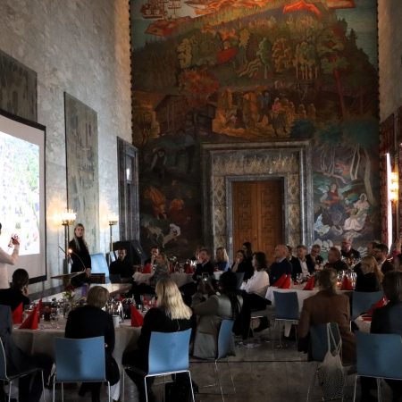 Folk sitter ved bordene i en storstue med et stort veggmaleri. En presentatør står ved en skjerm som viser en presentasjon. Rommet lyses opp av flere vinduer til høyre.