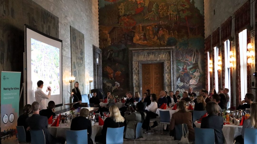 En person presenterer en lysbildefremvisning for et publikum i en utsmykket dekorert hall med veggmalerier.