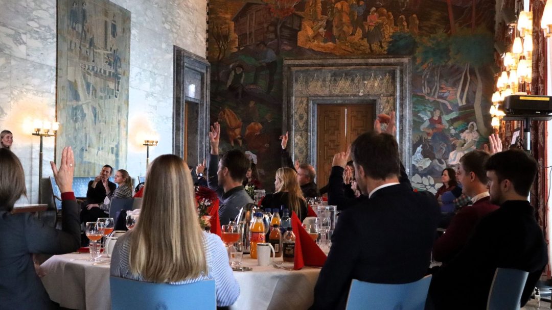 Folk satt rundt bordene i et stort rom og rekker opp hendene. Vegger utsmykket med billedvev og kunstverk, stemningsbelysning til stede.