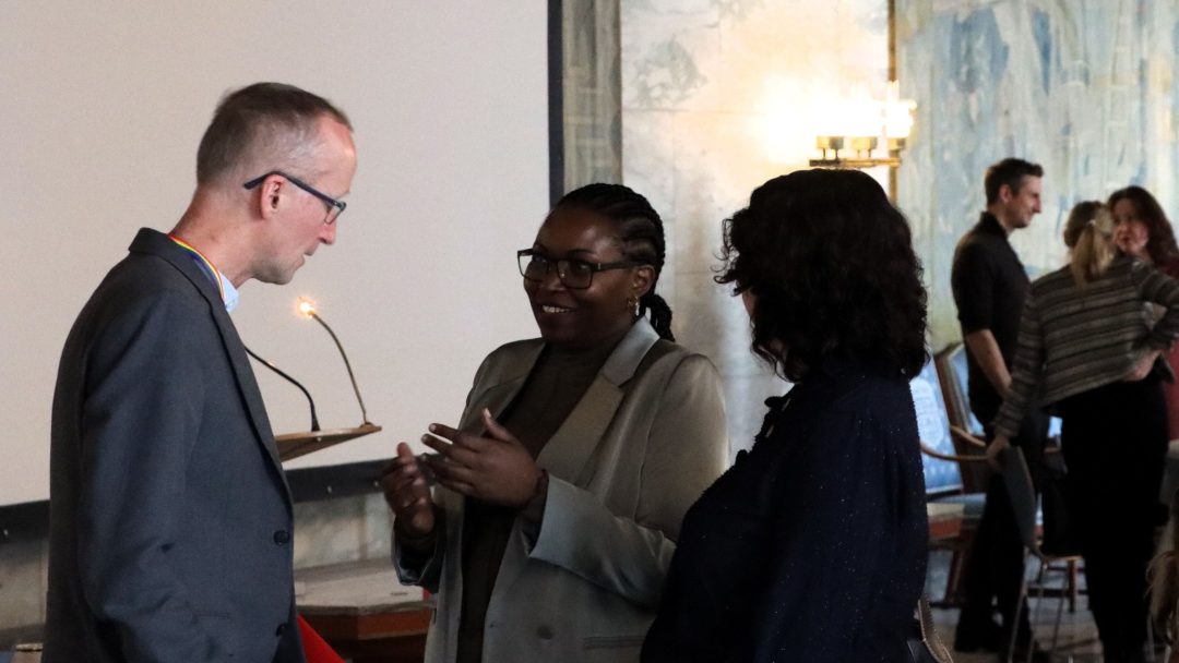 Tre personer gikk i samtale i innendørs omgivelser med et podium og andre i bakgrunnen.