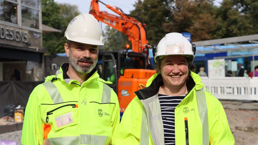 To bygningsarbeidere iført knallgule vernejakker og hjelmer står foran en oransje gravemaskin på en utslippsfri byggings byggeplass.