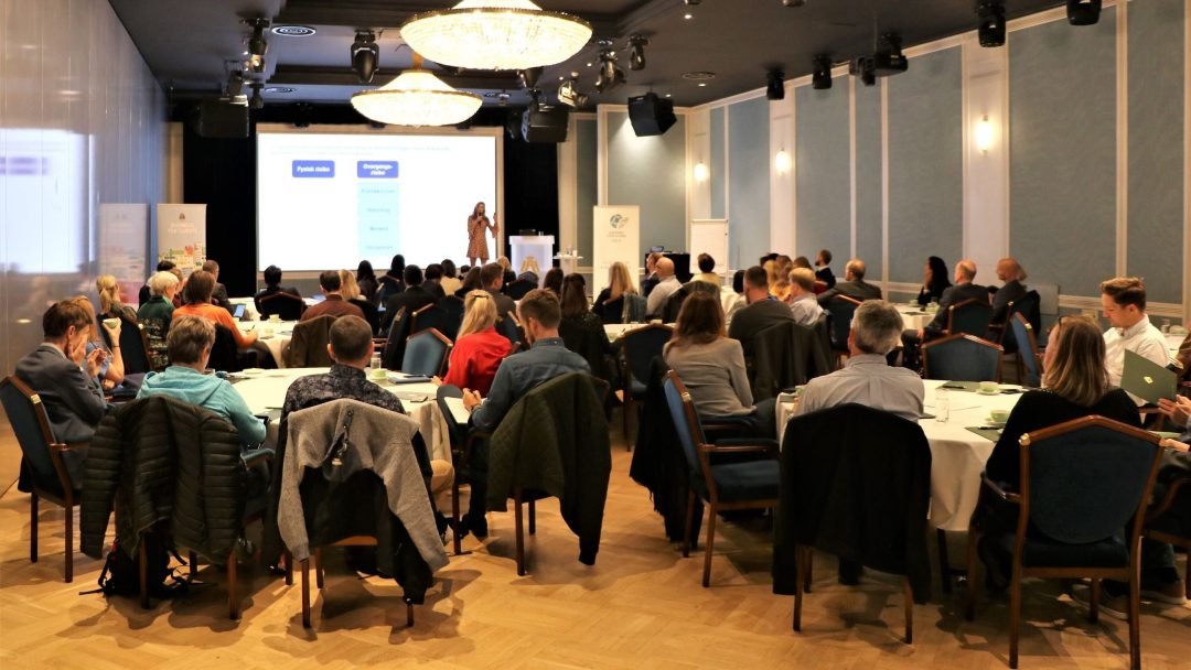 En foredragsholder presenterer for et publikum som sitter ved runde bord i et konferanserom med lysekroner.