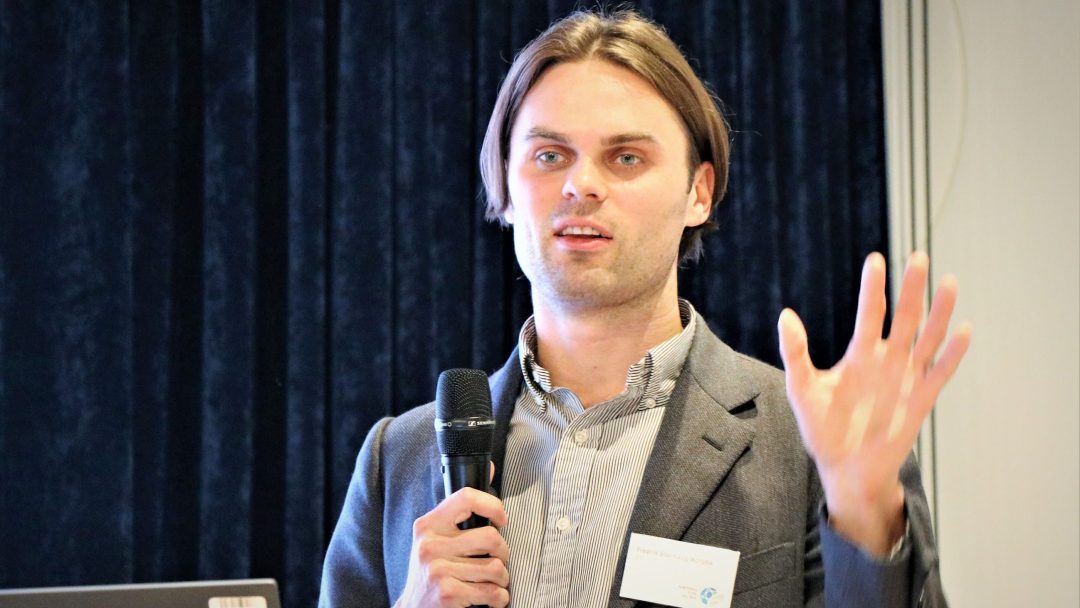 En person i dress gestikulerer mens han snakker i en mikrofon foran et mørkt gardin.