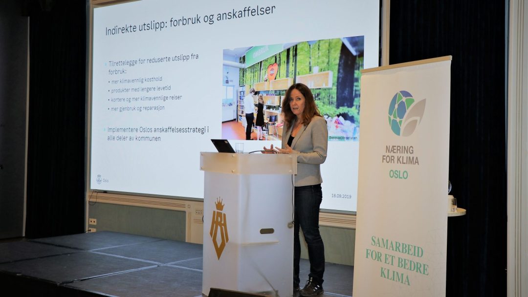 En kvinne står på et podium og holder en presentasjon om indirekte utslipp. En skjerm med kulepunkter er bak henne, og et banner med klimatema er ved siden av podiet.