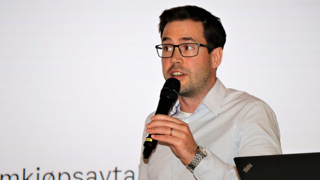 En person med briller og hvit skjorte snakker i en mikrofon, stående foran en projeksjonsskjerm.