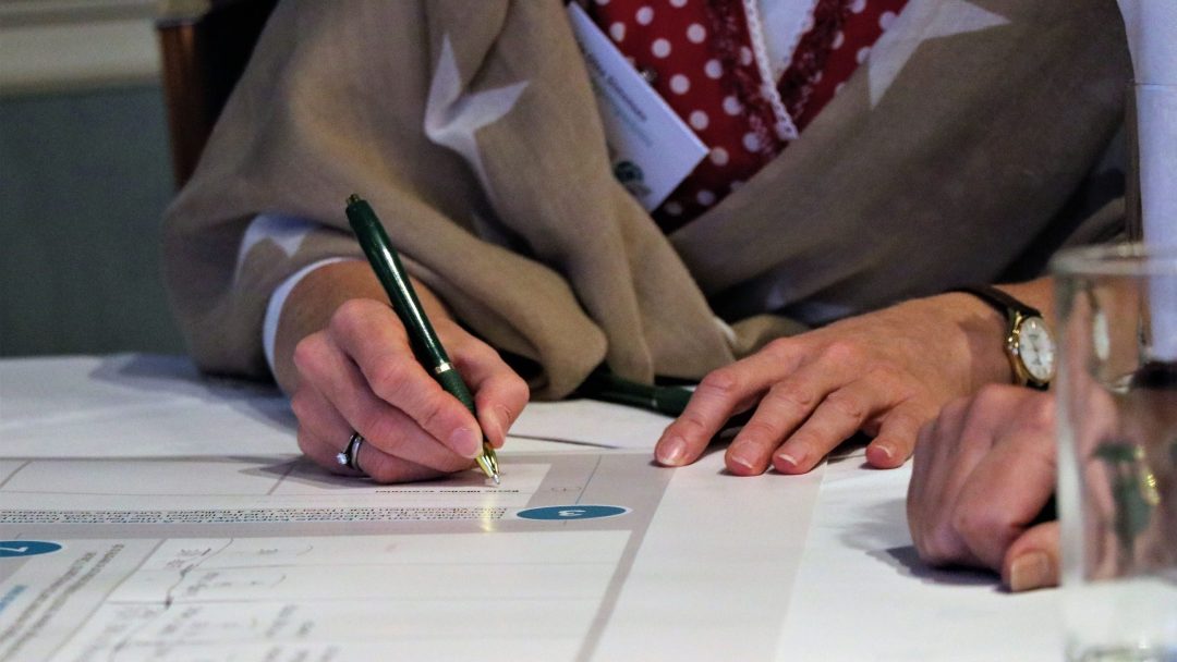 En person som har på seg et sjal, signerer et dokument med en grønn penn ved et bord.