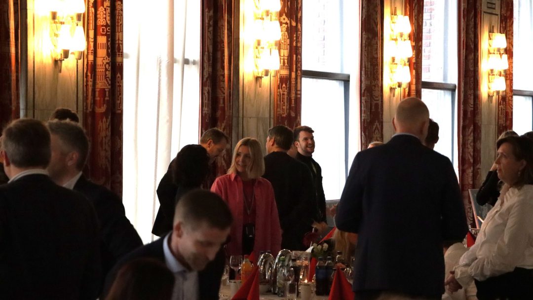 Folk som står og samhandler i et svakt opplyst rom med bord dekket til bespisning.