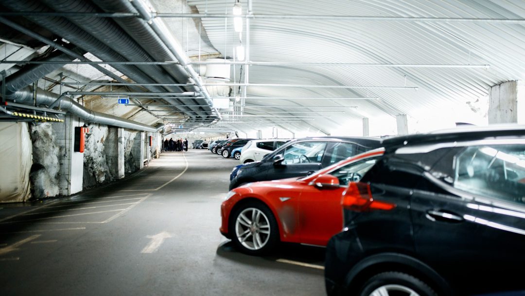 En underjordisk parkeringsplass med rader med parkerte biler, inkludert en fremtredende rød bil i forgrunnen. Det godt opplyste, romslige området gjenspeiler den økende trenden med elektrifisering med ladestasjoner plassert praktisk overalt.