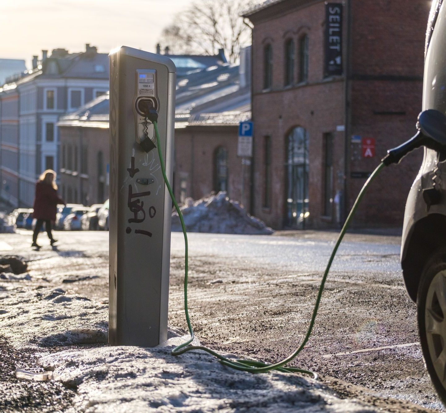 Elbil koblet til en ladestasjon i en snødekt gate i et urbant område, med bygninger og en gående person i bakgrunnen.