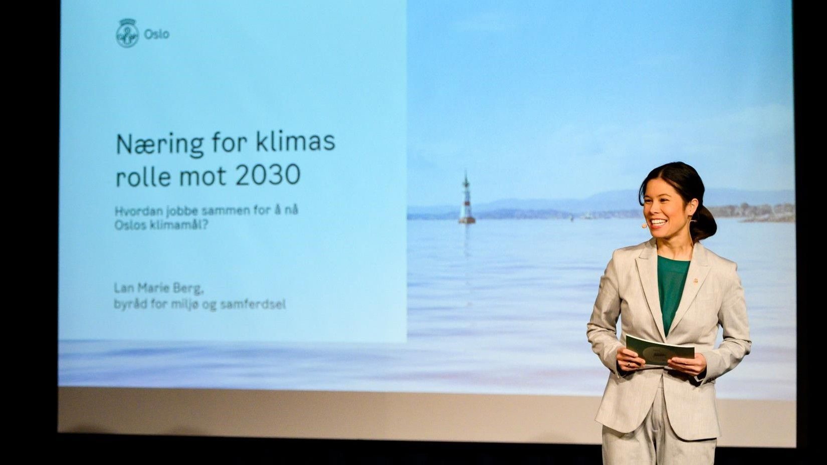 En kvinne står på scenen med papirer, foran en skjerm som viser en presentasjon om Oslos klimarolle frem mot 2030.