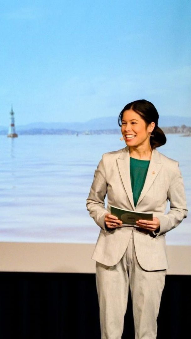 Person i lys grå dress står på scenen og holder papirer og smiler. Bakgrunnen viser en havscene med en bøye og fjerne åser.