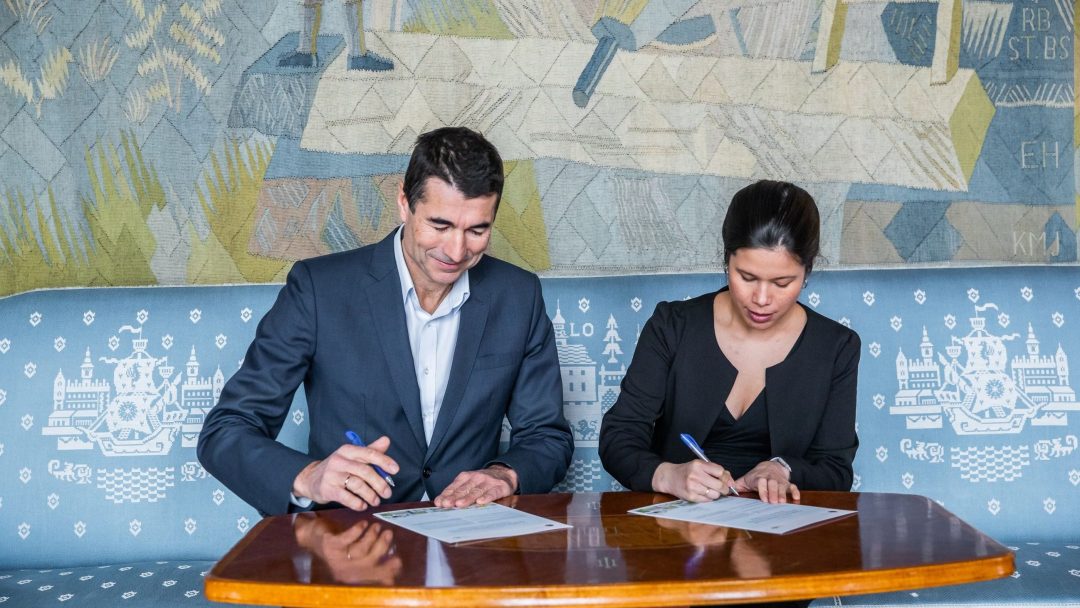 To personer sitter ved et bord og signerer dokumenter med penner. De er kledd i formell antrekk. Et dekorativt veggteppe er i bakgrunnen.