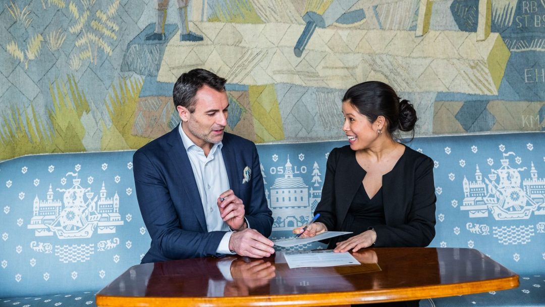 En mann og kvinne i formell antrekk sitter på en mønstret benk og diskuterer dokumenter ved et trebord med et veggmaleri i bakgrunnen.