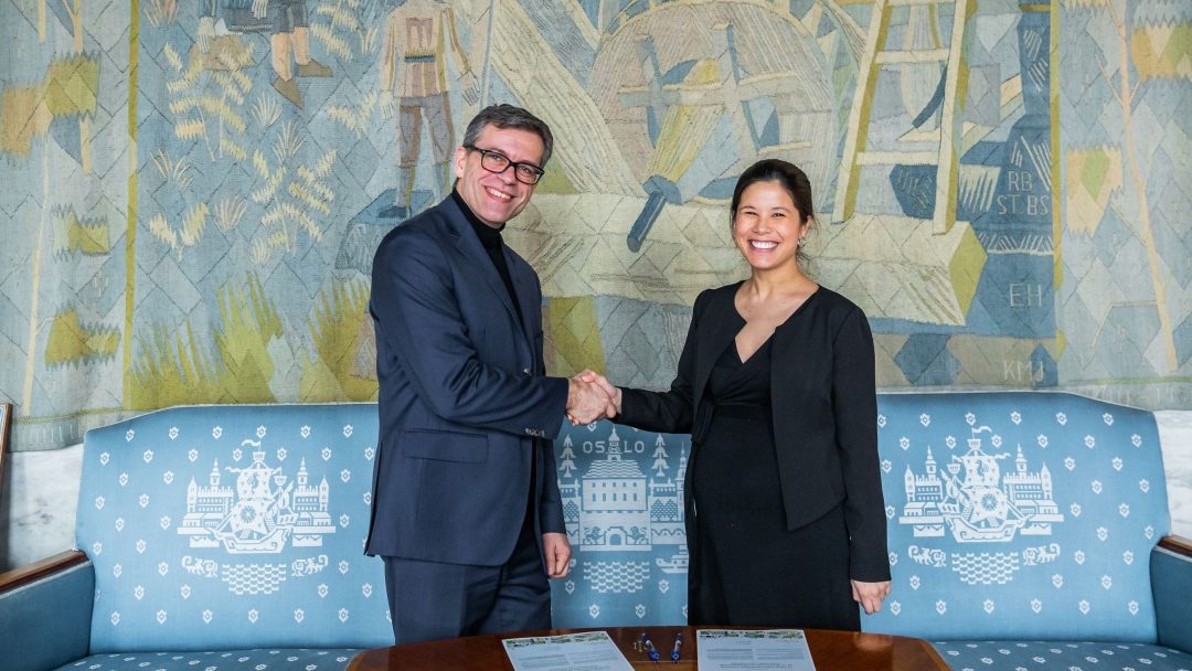 To personer håndhilser foran et dekorativt billedvev. De smiler, har på seg formell antrekk og står i nærheten av en blå sofa med mønstre med Oslo-tema. Dokumenter og penner ligger på bordet.