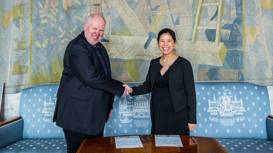 To personer i formell antrekk håndhilser på et bord med papirarbeid, foran et dekorativt veggteppe. Begge smiler.