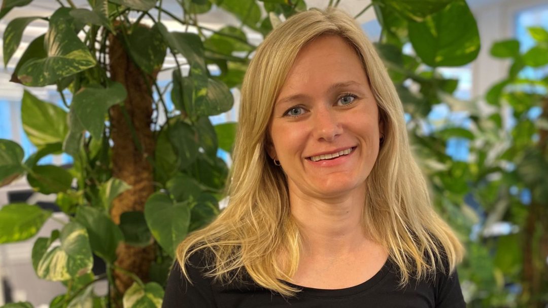 En person med blondt hår smiler mens han står blant frodige grønne planter innendørs, og legger til et snev av natur til deres rolige scene.