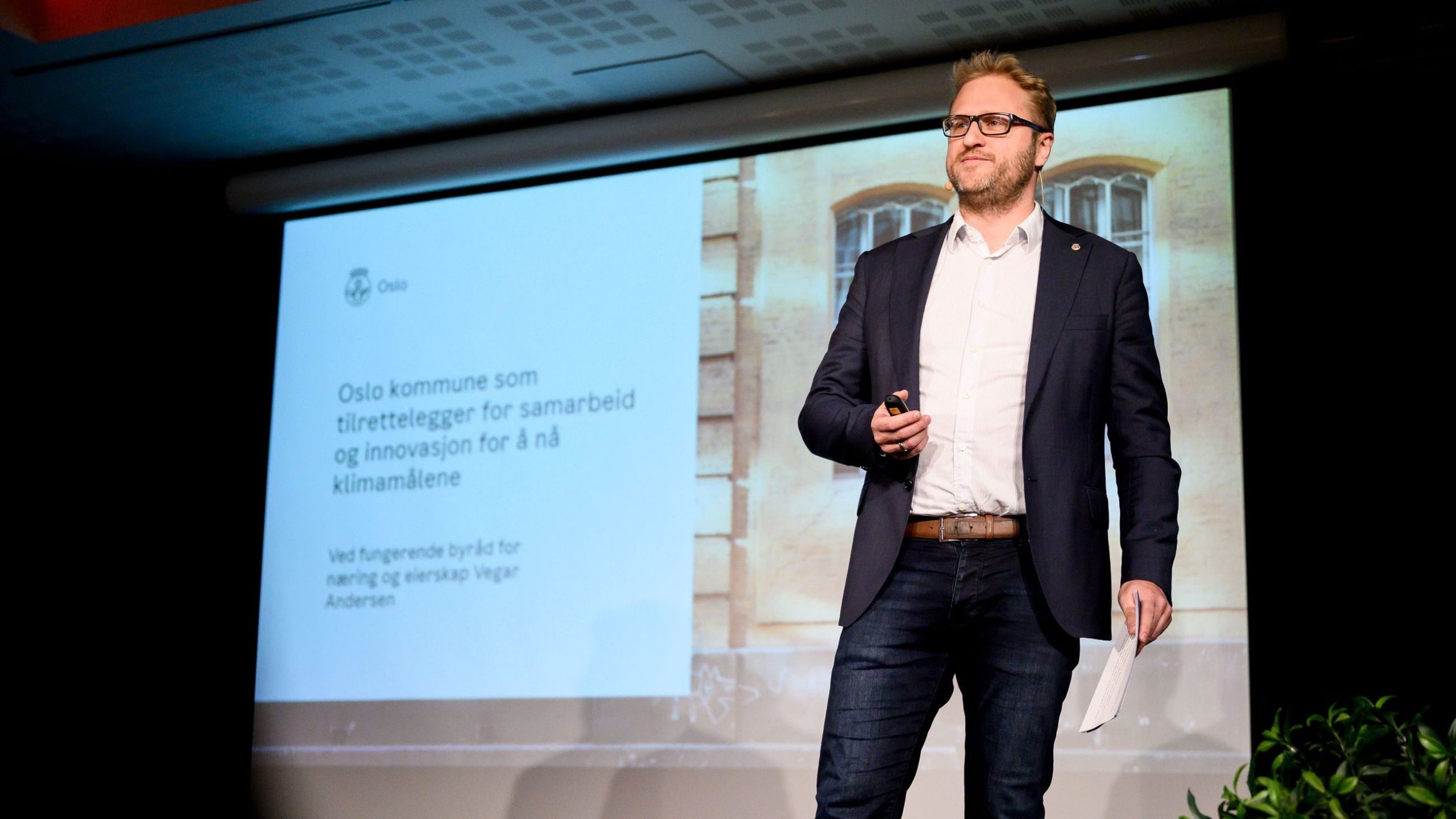 En mann i dress holder en mikrofon på scenen med en presentasjon projisert bak seg om Oslos klimamål og innovasjoner.