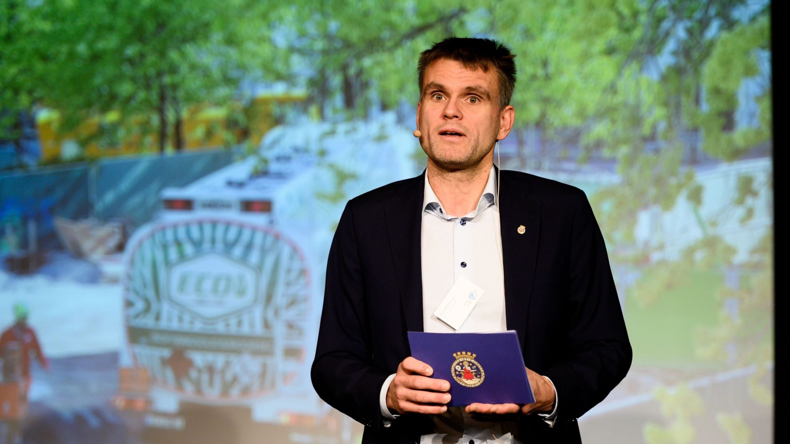 En mann i dress og hvit skjorte holder en blå mappe mens han står foran en skjerm som viser en naturscene.