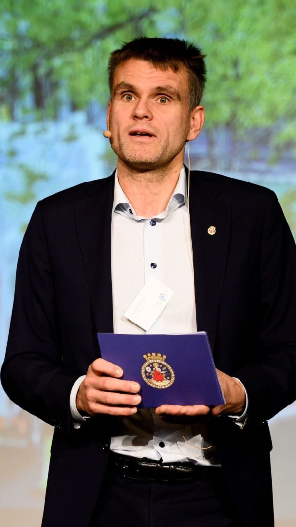 Mann i dress snakker på scenen, holder en blå mappe med et emblem.