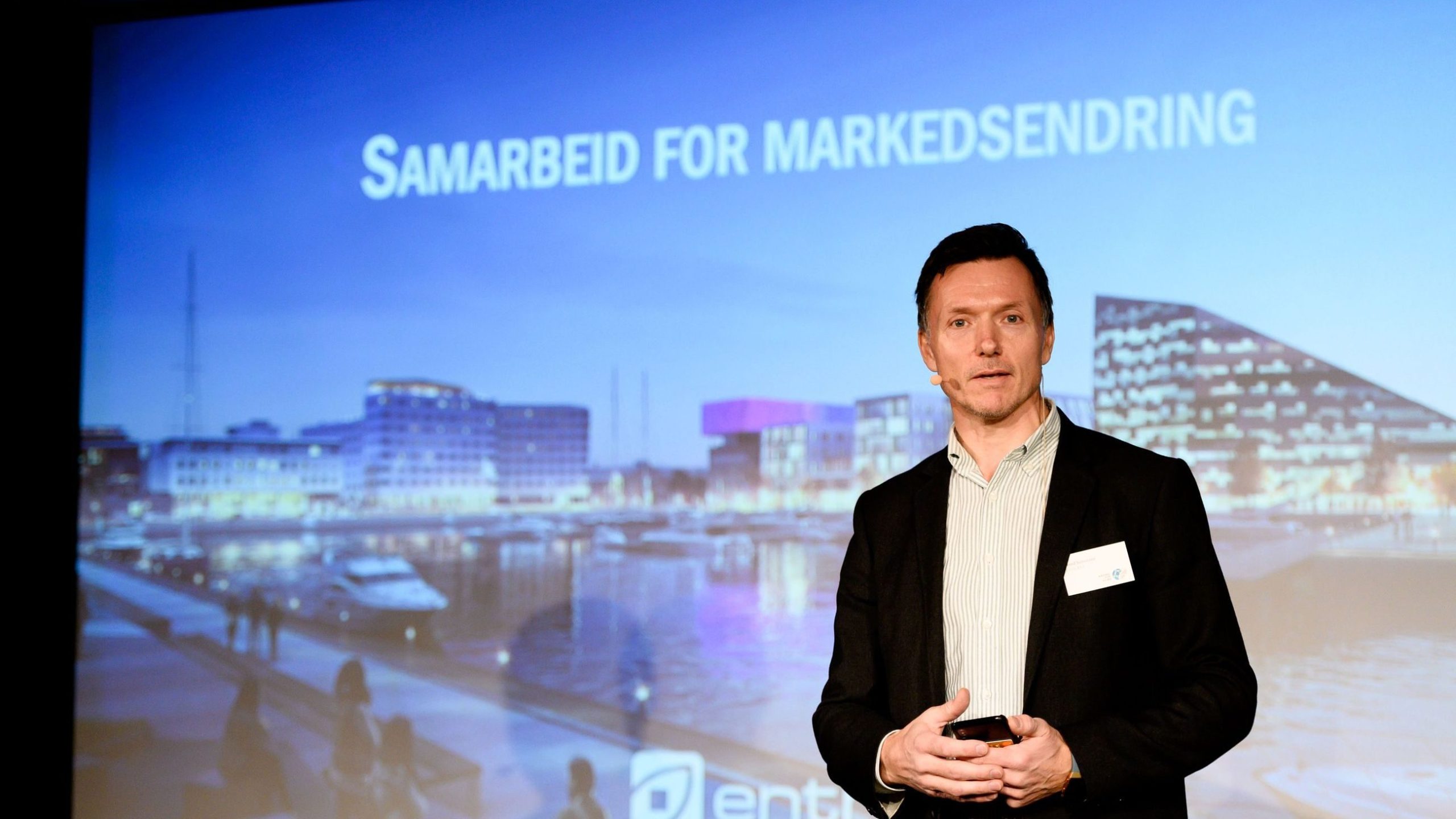 En mann står på scenen med et bybilde bak seg og tekst som lyder «Samarbeid for Markedsendring.
