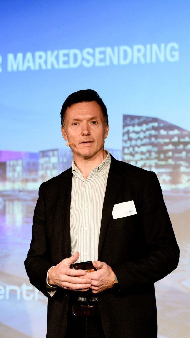 Person i dress som snakker på scenen med bybildebakgrunn.