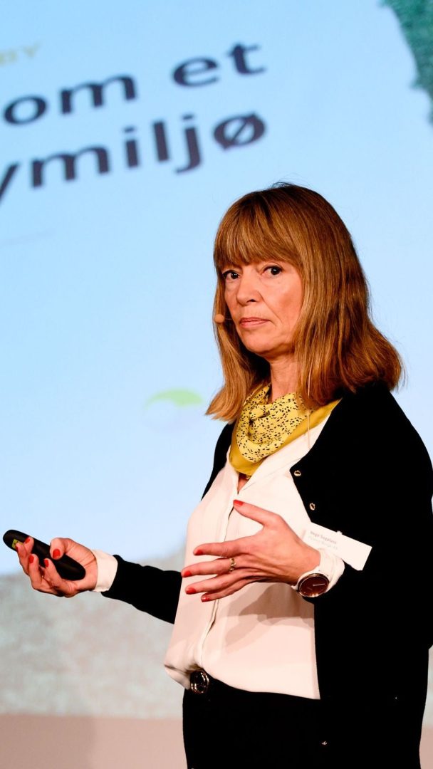 En kvinne som holder en mikrofon og presenterer foran en skjerm som viser tekst.