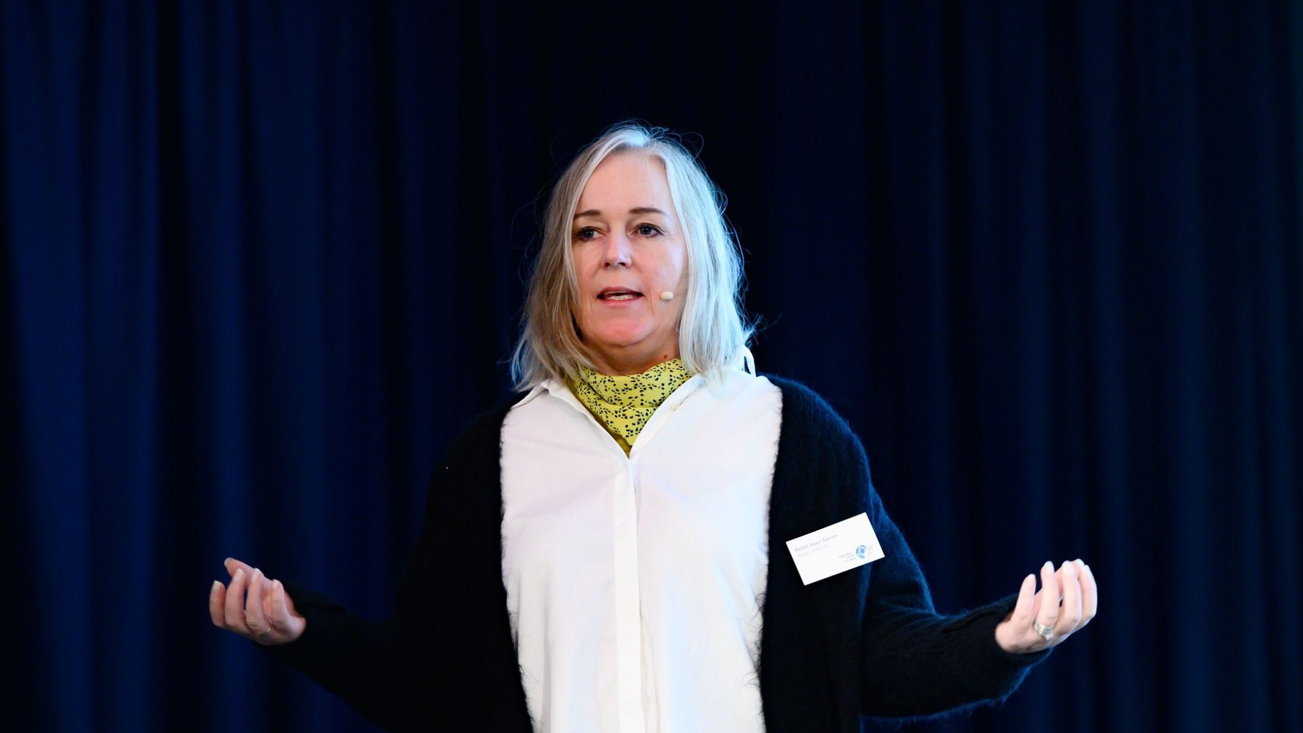 Person med hvitt hår som snakker på scenen, iført hvit skjorte og svart genser, med mikrofon på.