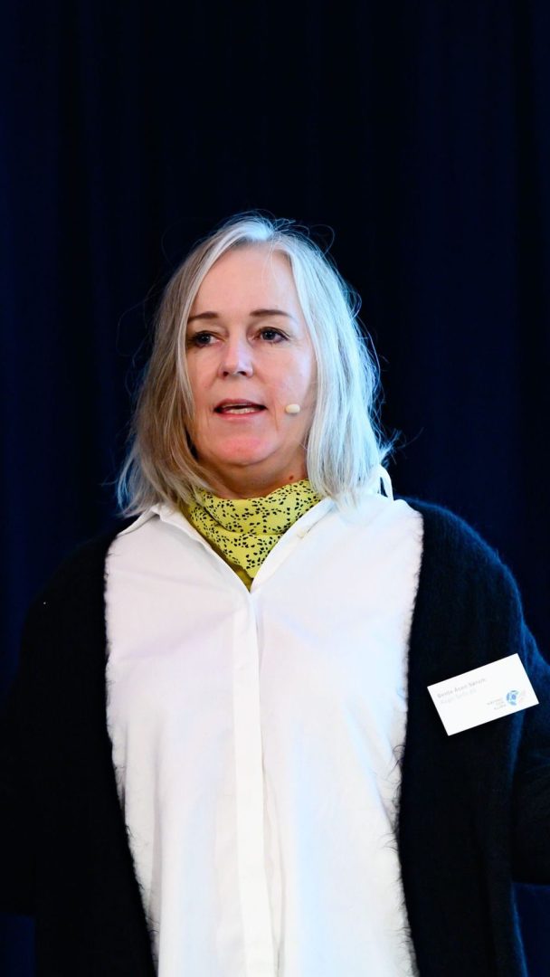 En kvinne med skulderlangt grått hår, iført hvit skjorte, svart cardigan og gult skjerf, snakker på scenen med et mikrofonhodesett.