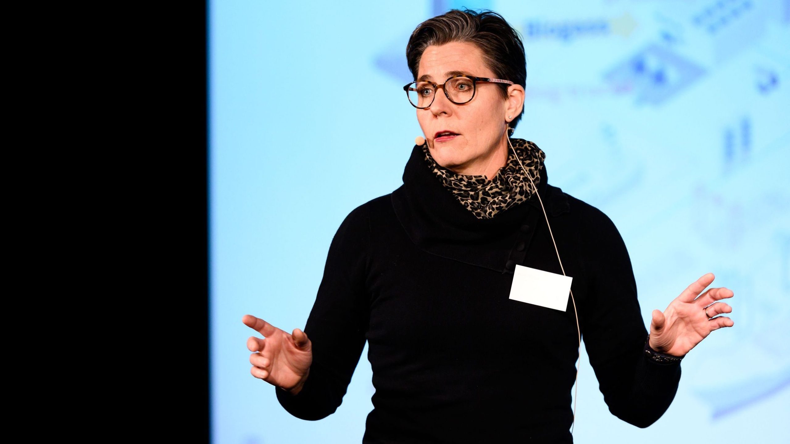 Person med kort hår og briller snakker, iført en svart topp og leopardtrykt skjerf, gestikulerer med hendene.