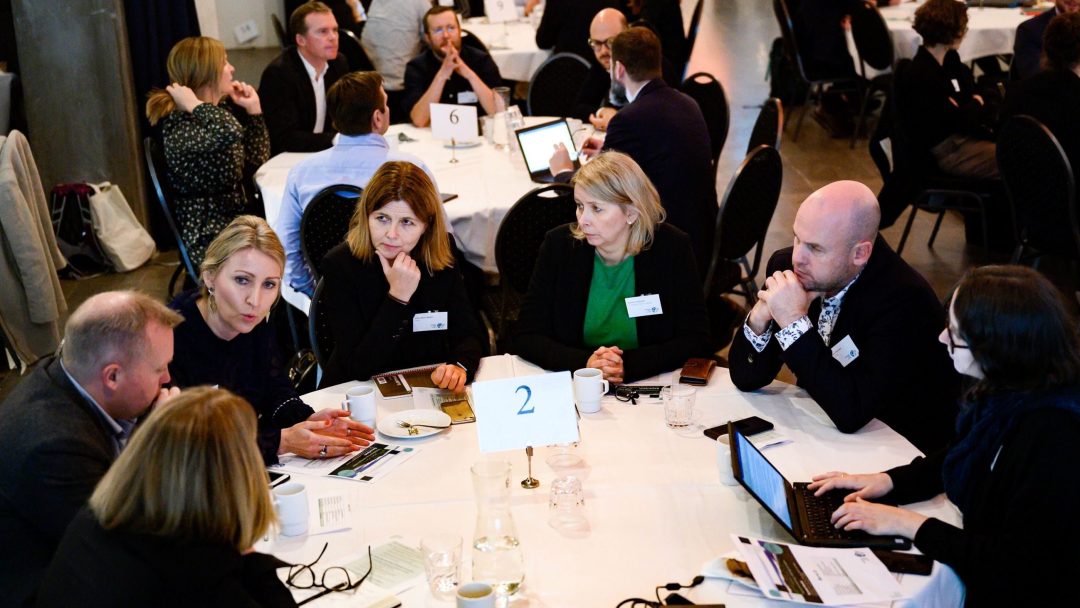 Folk sitter rundt et bord, engasjert i diskusjon, med bærbare datamaskiner og dokumenter. Et skilt med tallet 2 er på bordet.