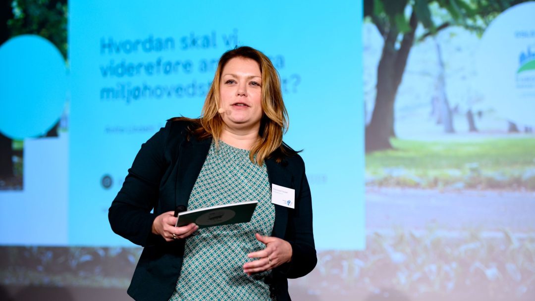 Kvinne snakker på scenen med et presentasjonslysbilde i bakgrunnen. Hun holder en mappe og har på seg en blågrønn mønstret kjole med en mørk blazer.